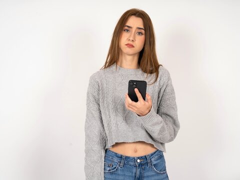 Upset dissatisfied Caucasian young girl wearing grey sweater uses mobile software application and surfs information in internet, holds modern mobile hand