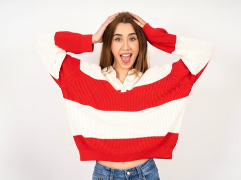 Cheerful overjoyed Caucasian young girl wearing striped sweater reacts rising hands over head after receiving great news.