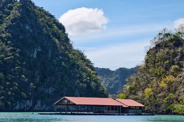 Floating Wooden House Between Jungle-Clad Limestone Hills on Tranquil Tropical Lake Under Blue Sky
