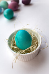 A green and blue Easter egg stands in a white porcelain tartlet with straw on a white background