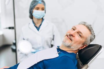 Obraz premium Smiling mature male patient during consultation, female dentist in medical gloves holding mirror