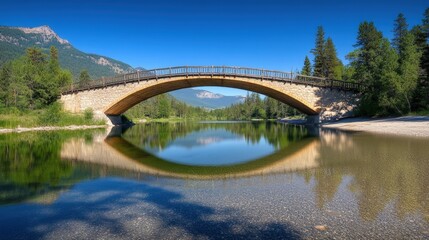 Fototapeta premium A serene landscape featuring a stone bridge over a calm river, surrounded by lush greenery and distant mountains under a clear blue sky.
