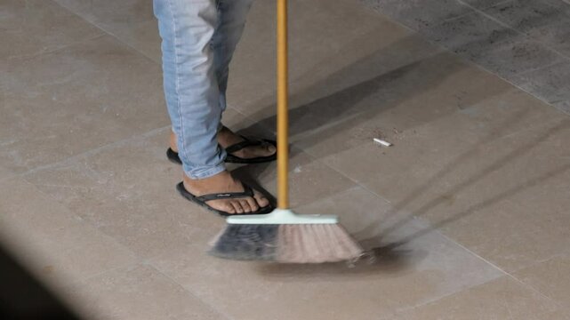 Man in jeans and sandals with broom brush sweeps away trash cigarette butt from sidewalk road pavement tiles, at night, close up legs. Shop owner keeps area clean. Seller cleans up garbage, tool