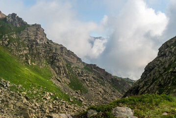 High mountain scenery with partly rocky hill with steep slopes and higher mountain peaks on the background mostly covered by clouds