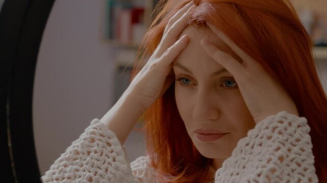 Woman with long red hair feeling strong headache and migraine touching her head and forehead in front of a mirror