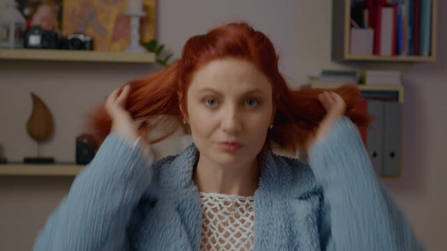 Young woman with red hair puts on a blue coat and thoughtfully checks her reflection in the mirror to ensure she hasn't forgotten anything. After a moment, she gathers her things and heads out.