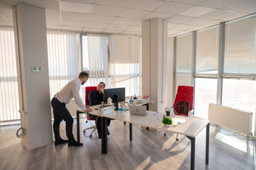 Caucasian man standing at colleague's desk discussing work. 
