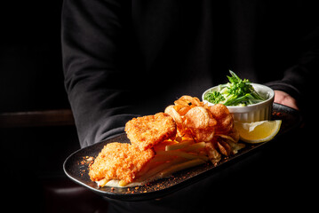 A server holds a platter featuring crispy fried fish fillets with golden fries, served alongside fresh salad and a slice of lemon, elegantly arranged on a dark rectangular plate.