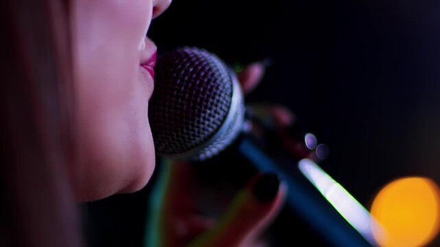 Beautiful girl singer sings into a microphone on stage against the background of multi-colored lights, close-up. Painted lips and microphone, slow motion