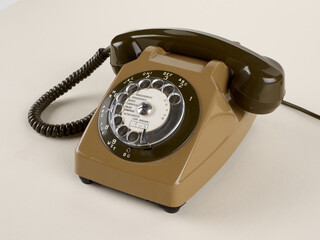 Un téléphone à cadran rotatif de couleur marron avec ses câbles. Des chiffres et des lettres pour la numérotation sont visibles. Sur fond beige.