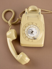 Un téléphone à cadran rotatif de couleur beige, avec écouteur secondaire, avec ses câbles. Des chiffres et des lettres pour la numérotation sont visibles. Sur fond marron.
