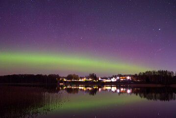 Northern lights glow green and purple over a calm lake at night, reflecting stars and trees in the water.