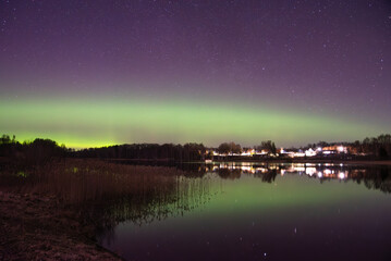 Bright green northern lights dance above a village at night, reflecting in the lake under a star-filled purple sky.