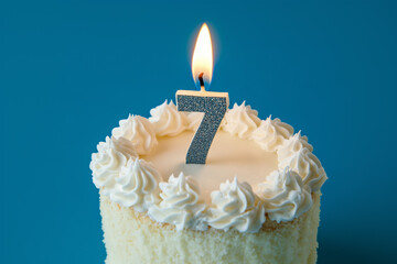 Lit candle in shape of number 7 on delicious happy birthday cake dessert close up detail against pastel blue background.