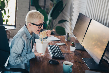 Young female programmer eating noodles while multitasking at her home office workspace