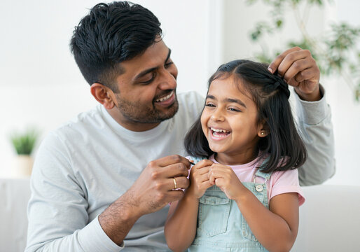 Happy indian father braiding his cheerful daughter's hair, enjoying a tender moment together in their living room