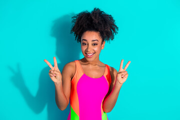 Bright athletic woman in vibrant sportswear showing peace gesture on a turquoise background with...