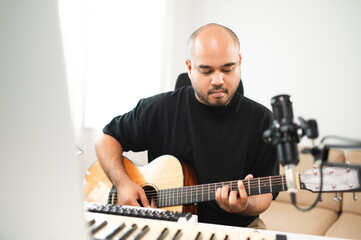 Asian male teenager playing guitar, composing music in home recording studio using computer,...