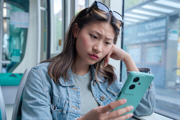 Young Chinese woman sitting by the window in public transport, reading her smartphone with a...