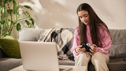 Beautiful teenage girl holding film camera and listening to tutorial on laptop while exploring basics of photography at home. Concept of education, homeschooling, studying, creative hobby