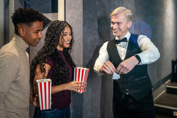 Couple holding popcorn buckets entering movie theater, checking tickets with usher
