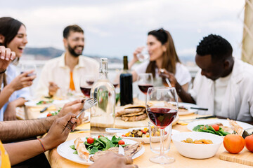 Happy group of multiracial young friends having dinner at rooftop summer party. Multiracial people...