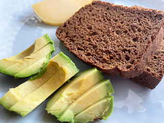 Two square toasts of black bread, sliced ​​green ripe avocado and pieces of hard cheese lie on a round gray textured plate, close-up, top view. Proper nutrition. Vitamins. Healthy snack.