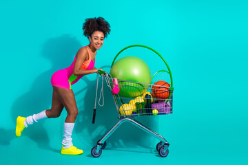 Young active woman in a vibrant bodysuit stands pushing a cart full of colorful fitness equipment