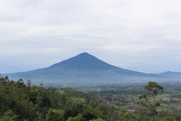 Fototapeta premium A majestic, solitary volcano dominates a hazy landscape, viewed from a distance. Lush green vegetation fills the foreground.