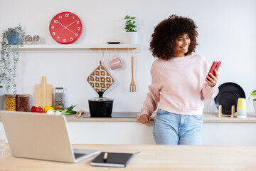 Young happy woman using mobile phone while telecommuting on laptop and cooking in modern kitchen. Business and technology concept
