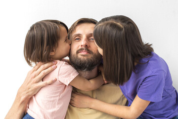 Fototapeta premium Two little daughters kissing bearded young father on cheeks while hugging him against white background, concept of family, love, parenthood