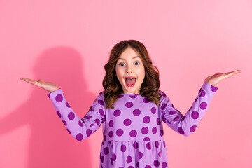 Happy cute girl with surprised expression wearing a purple polka dot dress with arms raised on a pink background © deagreez