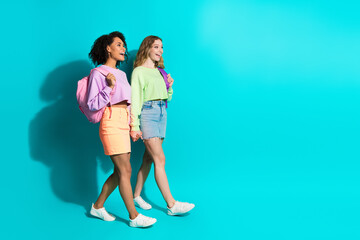 Fototapeta premium Two stylish young women walking confidently on a turquoise backdrop in vibrant apparel celebrating youth and friendship