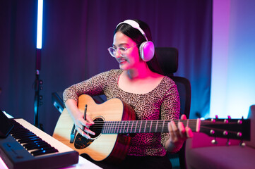 Asian teen girl playing guitar, composing music in home recording studio, LED lighting, at night using computer, headphones, condenser microphone, tablet and keyboard for mixing and mastering music.