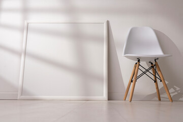Mockup of empty frame in modern minimalist interior with chair and morning light on white wall background. Template for artwork, painting, photo or poster