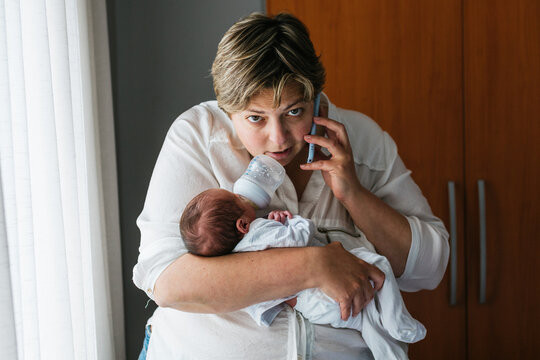 Mother holding newborn son and talking on phone while feeding him with baby bottle