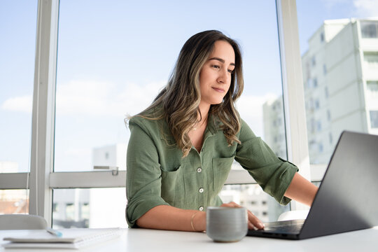 Focused Latin businesswoman in her 30s wearing green button-up shirt working on laptop with quiet contemplation in bright modern office with cityscape view and gray ceramic coffee mug on white desk