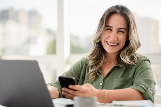 Cheerful Latin businesswoman in her 30s wearing green button-up shirt using smartphone while working on laptop in bright modern office with urban skyline view through large windows