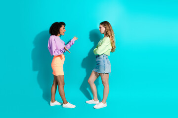Obraz premium Two young women discussing against a vibrant turquoise background, showcasing modern summer fashion and casual style with stylish outfits.