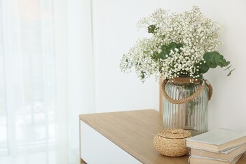 Beautiful gypsophila flowers with eucalyptus branches in vase and books on commode at home. Space...