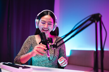 Teenager girl hosts podcast in her room using LED lights, computer, headphones, condenser microphone, tablet to take notes to share knowledge experiences in various fields via live online broadcast.