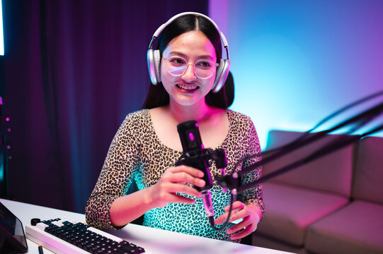 Teenager girl hosts podcast in her room using LED lights, computer, headphones, condenser microphone, tablet to take notes to share knowledge experiences in various fields via live online broadcast.