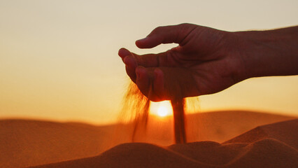 Young man takes a handful of sand from the desert and pours it against the background of the sunset with golden sunrays. Male hand touching a golden sand in the desert.