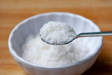 Close up of a spoon filled with coarse salt, held above a white bowl containing more salt