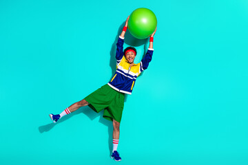 Energetic man lifting a large green exercise ball while wearing vibrant sporty attire against a solid light teal background