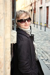 Outdoor portrait of adult woman in sunglasses with short blonde hair standing near a wall on empty street at springtime. Close-up of young female walking on the street and posing leaning against wall