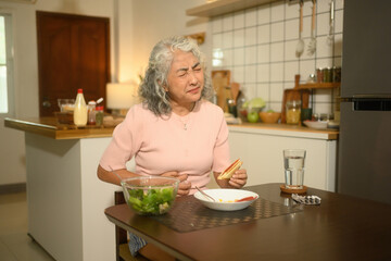 A senior woman in discomfort while eating breakfast, showing signs of digestive issues at home,...