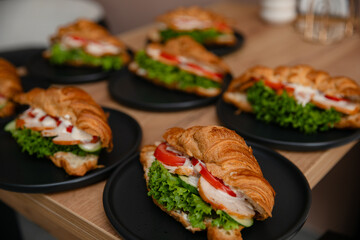 Delicious croissant sandwiches with lettuce, tomato, cucumber, chicken, and sauce on black plates, served on a wooden table. Perfect for breakfast, brunch, or catering presentations.