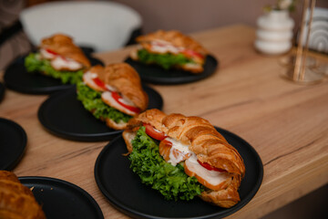 Delicious croissant sandwiches with lettuce, tomato, cucumber, chicken, and sauce on black plates, served on a wooden table. Perfect for breakfast, brunch, or catering presentations.