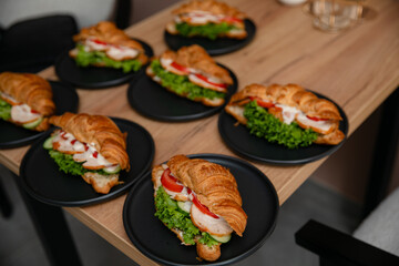 Delicious croissant sandwiches with lettuce, tomato, cucumber, chicken, and sauce on black plates, served on a wooden table. Perfect for breakfast, brunch, or catering presentations.